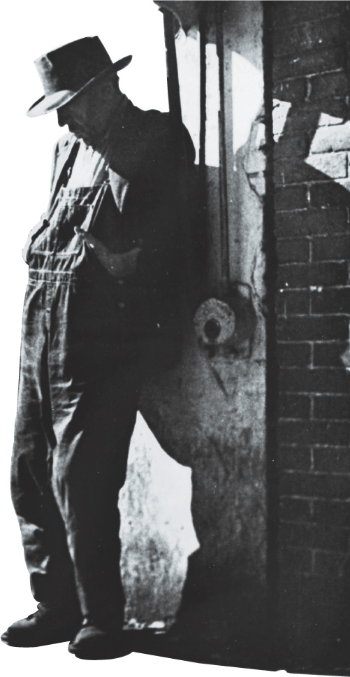 Photo: a man wearing overalls leans against a wall.