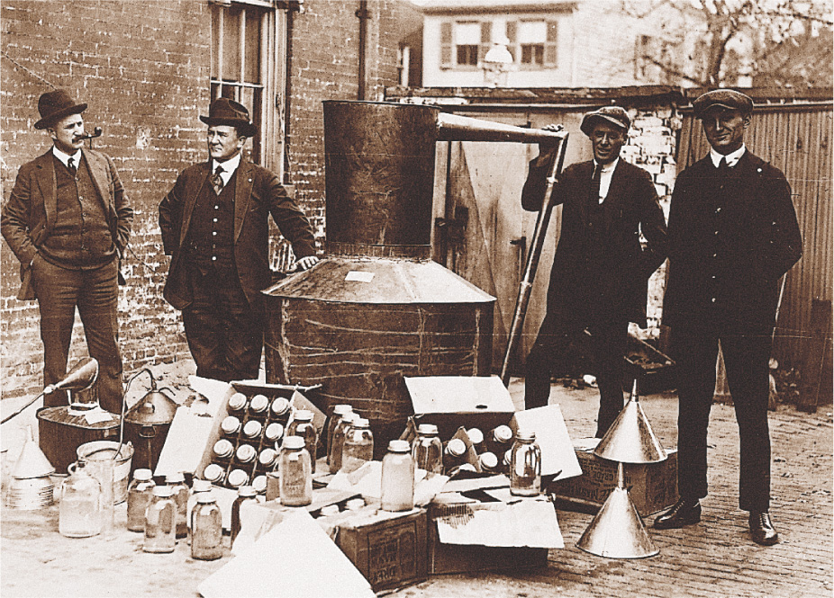 A photo: men stand by a metal distiller-tank and boxes of glass jars.