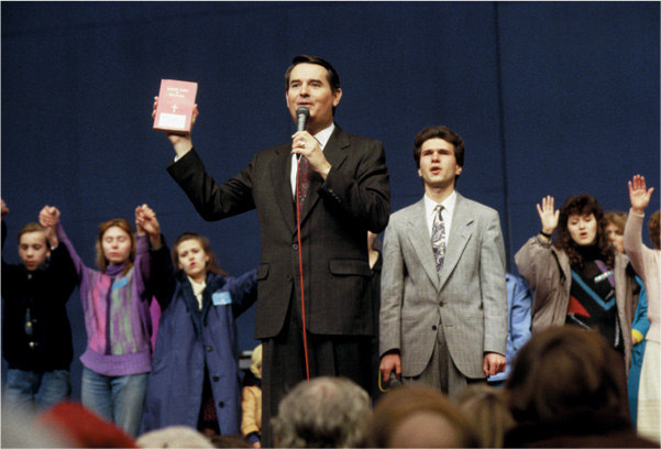 A photo: a
man in a suit holds a bible and speaks into a microphone. Behind him, people hold hands and raise
their arms.
