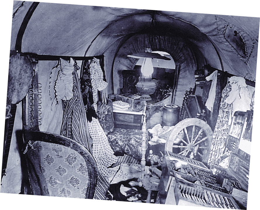 A photo: the interior of a covered wagon is packed full of clothes, furniture, a spinning wheel, and other household possessions.