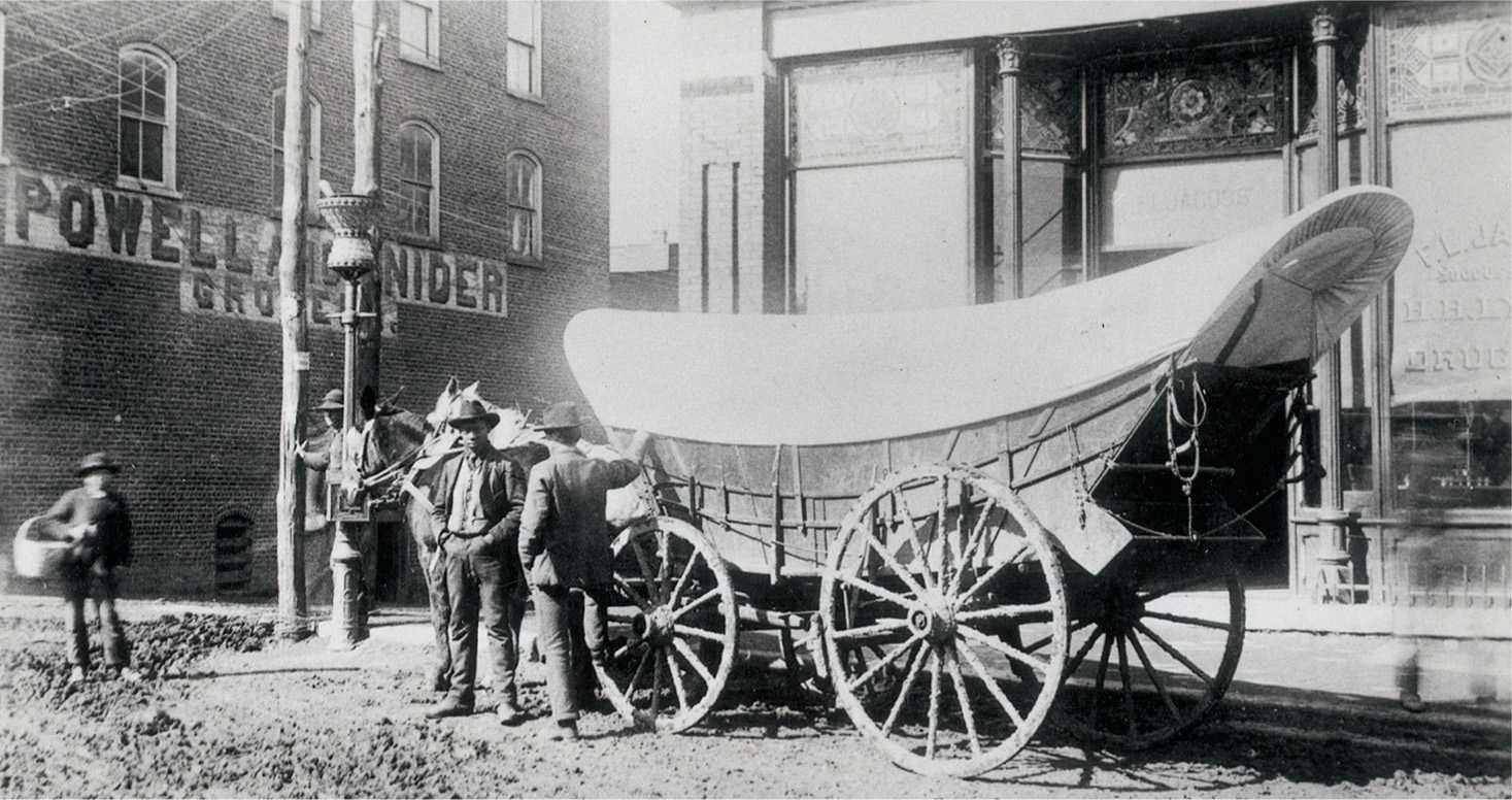A photo: on a dirt street in a town, two horses are hitched to a Conestoga wagon.