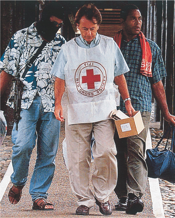 A photo: A man wears a smock labled Fiji Red Cross Society. Walking with him is another man wearing a ski mask, with a machine gun slung over his shoulder.