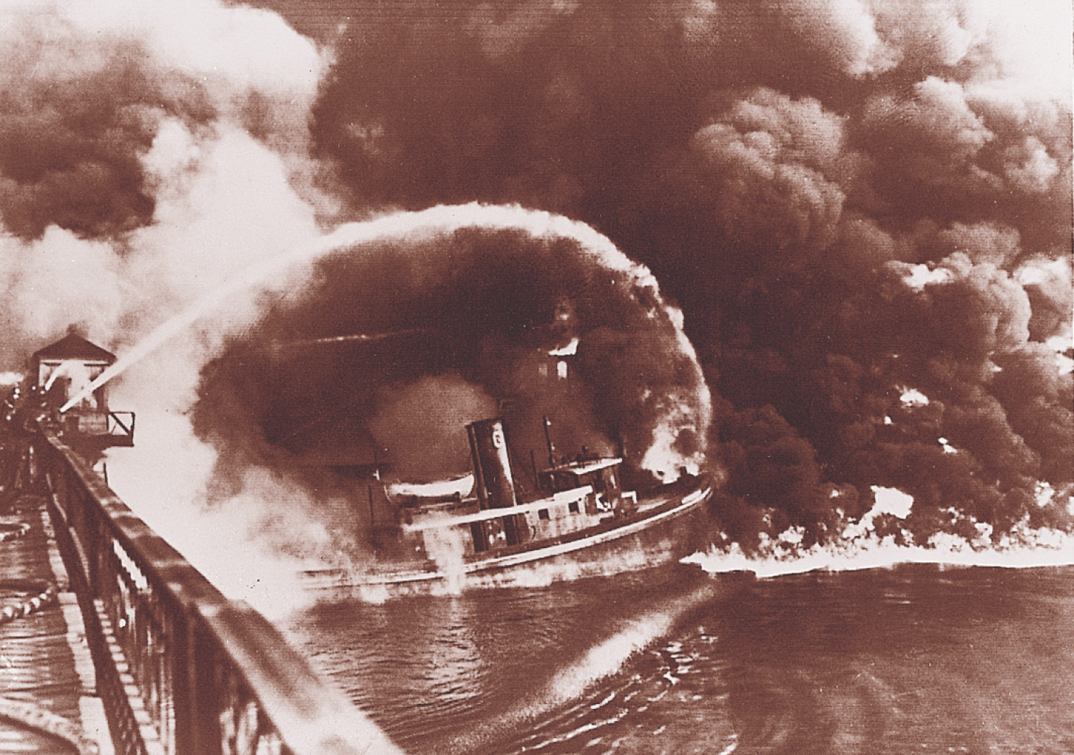 A photo: a tugboat sails toward flames burning on a river. Fireman spray water on the fire, under a sky filled with black clouds.