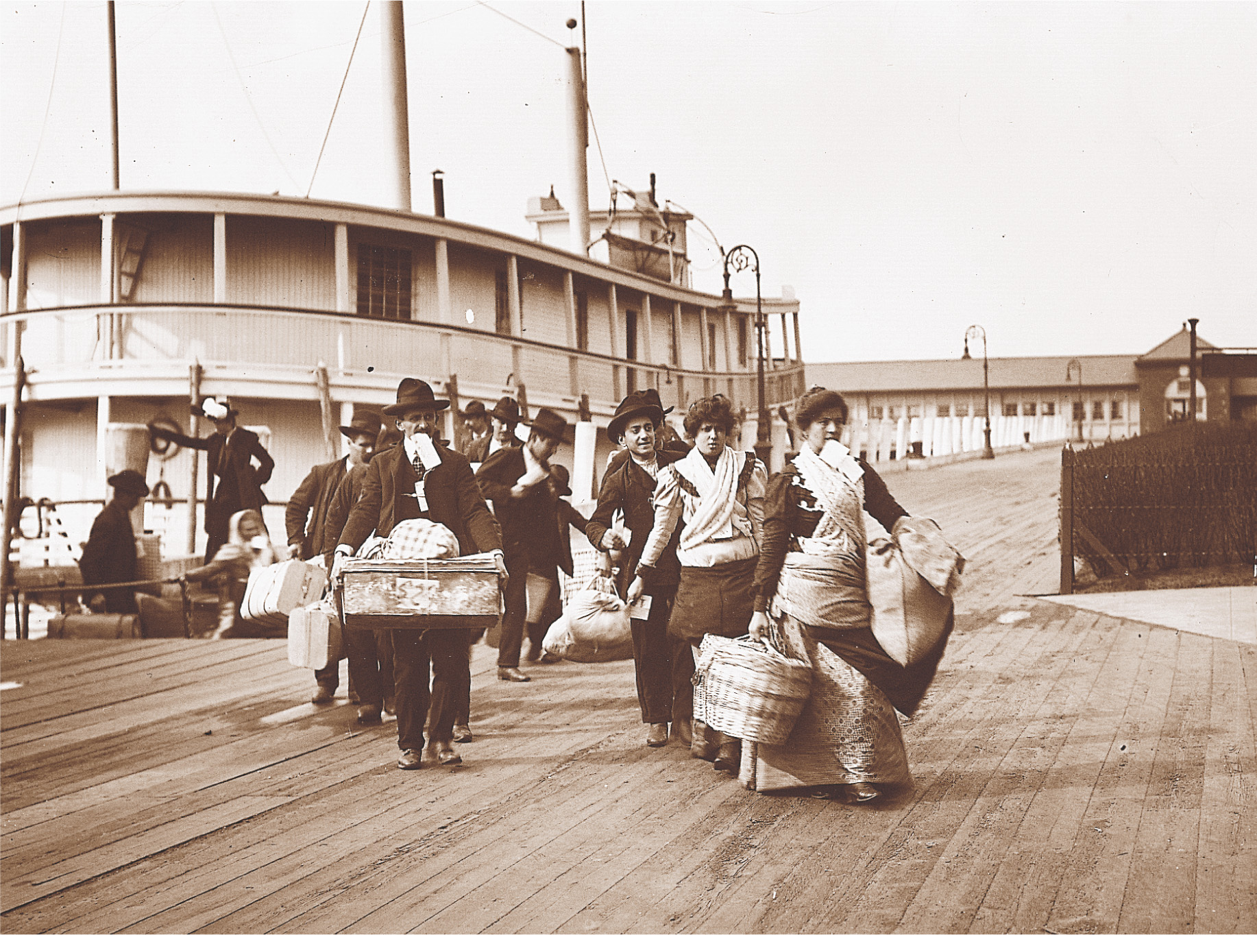 A photo: immigrants carrying suitcases step off a boat onto a dock.