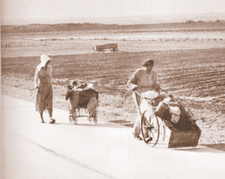 Photo: a couple pushes belongings in carts along a road next to a field