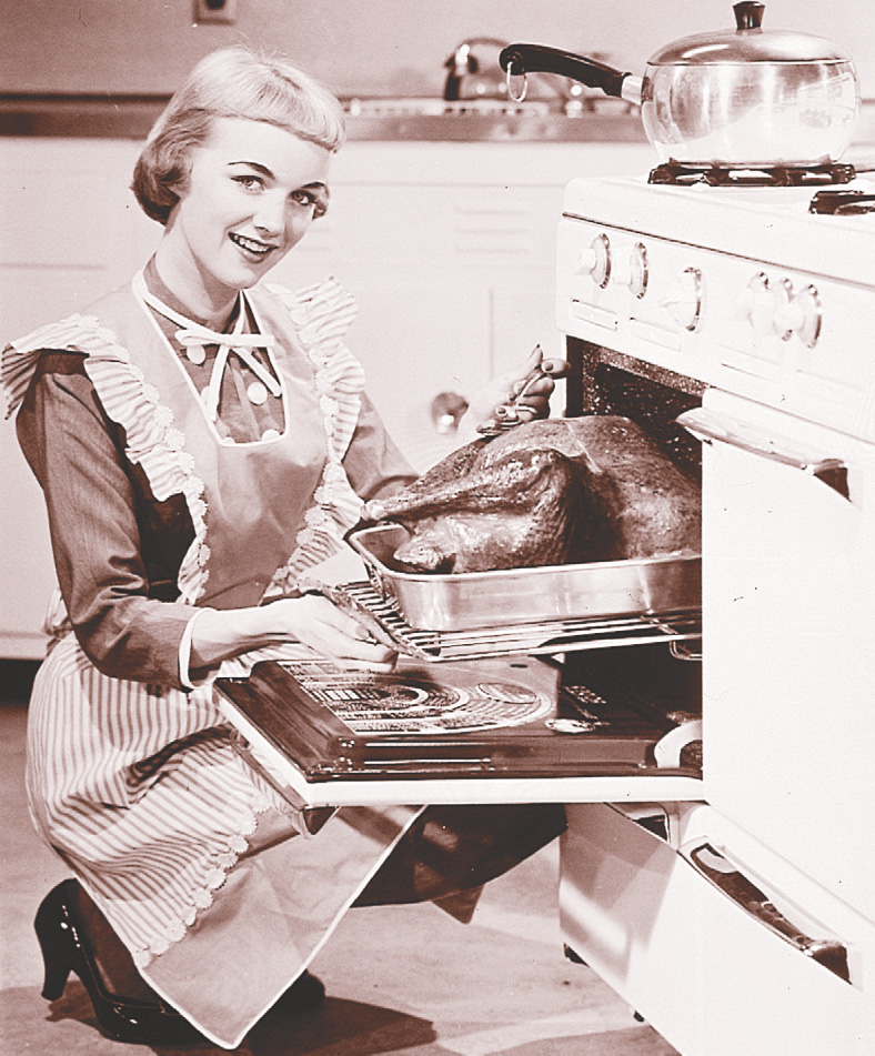 Photo: a smiling young woman wearing an apron removes a turkey from an oven