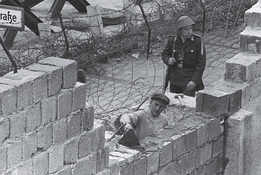 Photo: an armed soldier stands by a mason, who trowels cement over bricks in the unfinished wall.