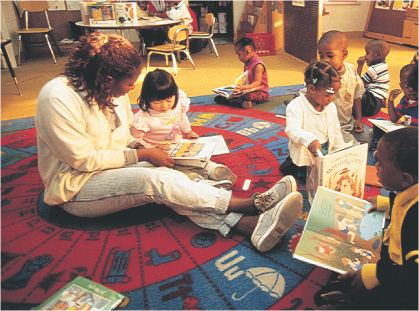 photo: teachers and young children sit on a classroom floor holding books.