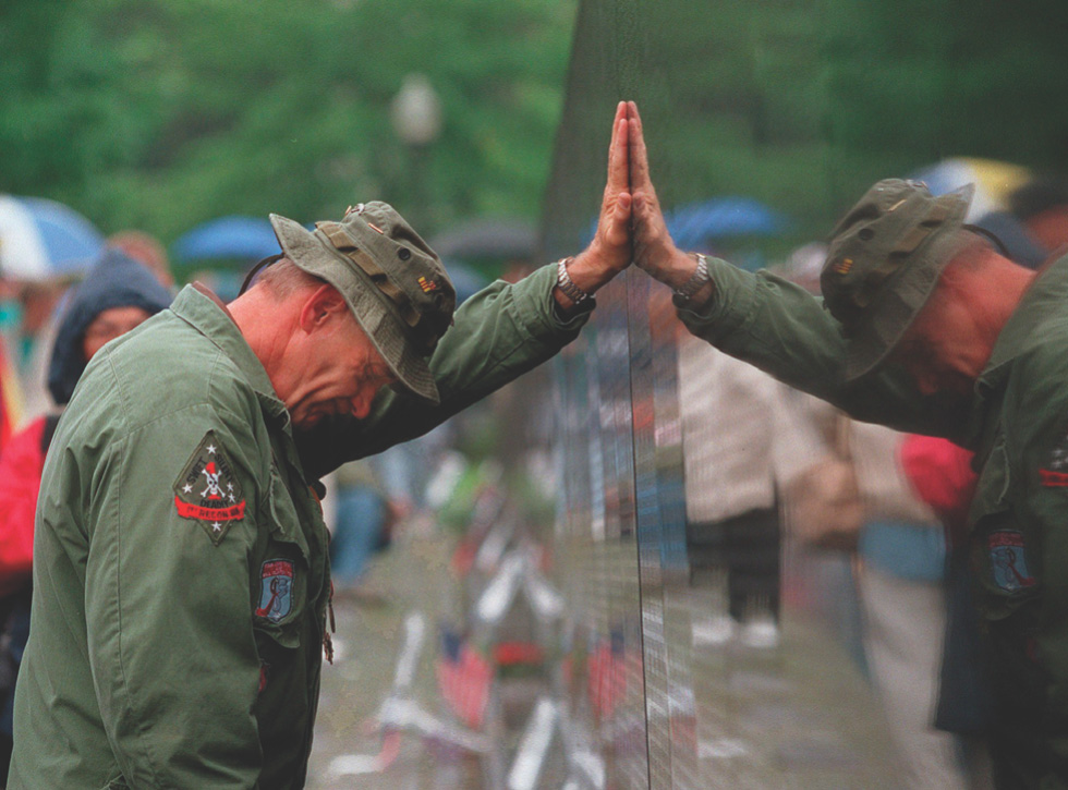 Photo: a man in a military coat leans forward and rests his hand on the Vietnam Veteran's Memorial wall, his head down.