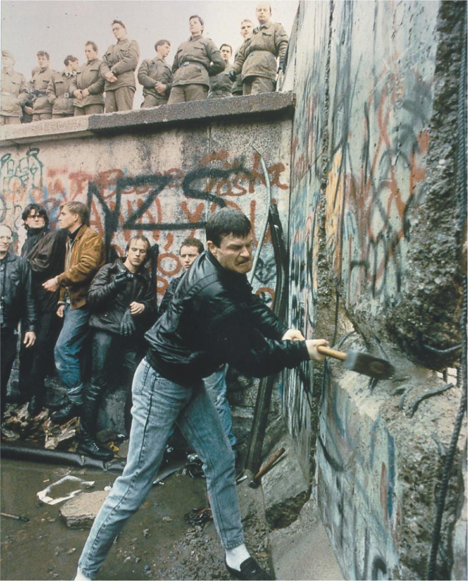photo: a man swins a sledgehammer at the graffiti-covered Berlin Wall.