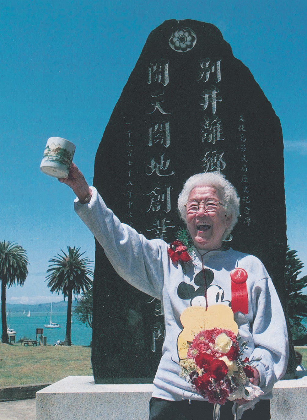 Photo: a white-haired Asian woman raises a mug in front of a monument.