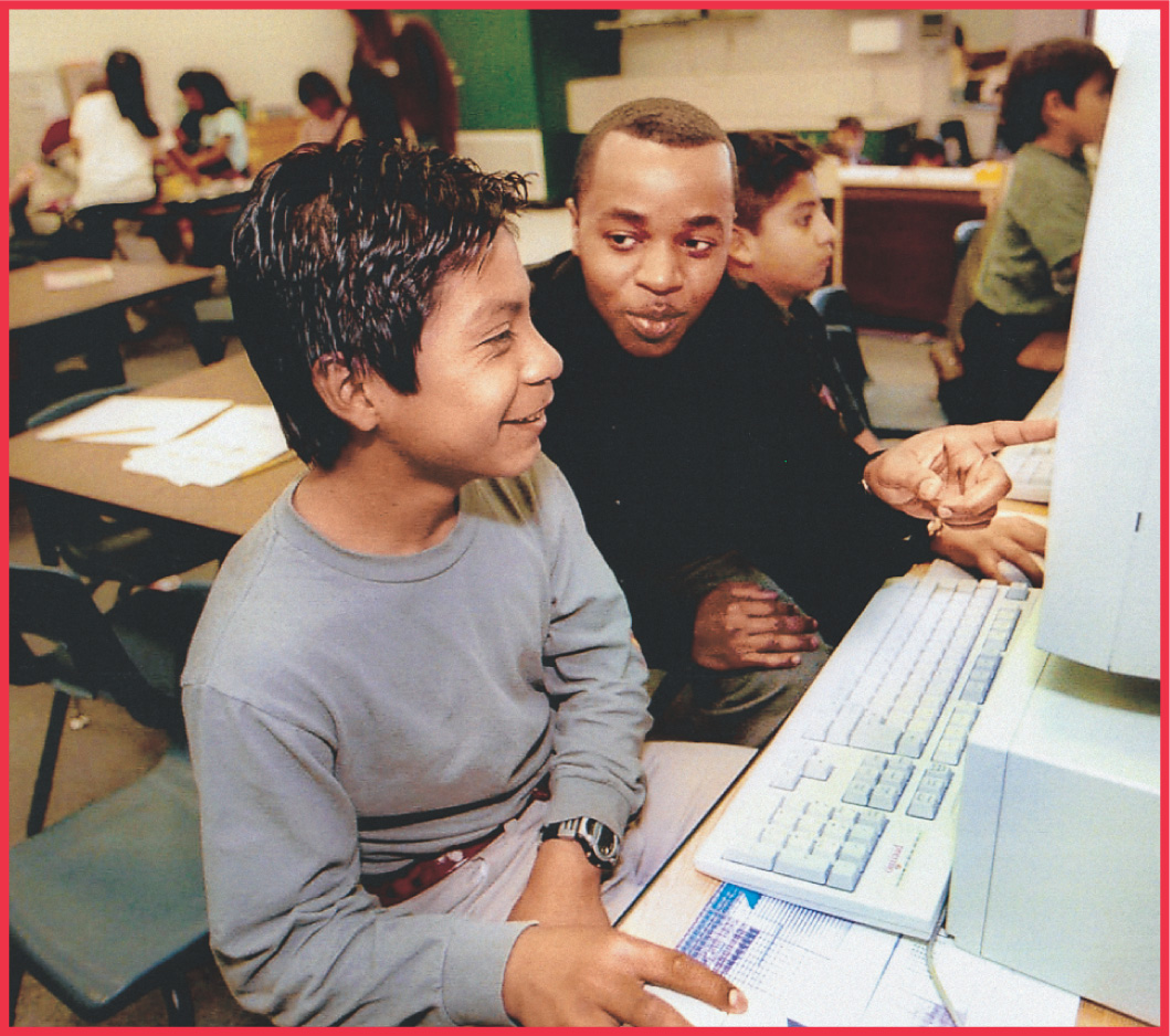 photo: a teacher helps a boy use a computer.